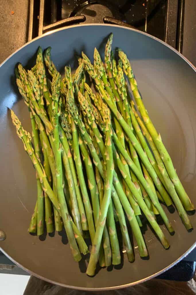 Fresh asparagus spears arranged in a single layer in a nonstick frying pan on a stovetop.