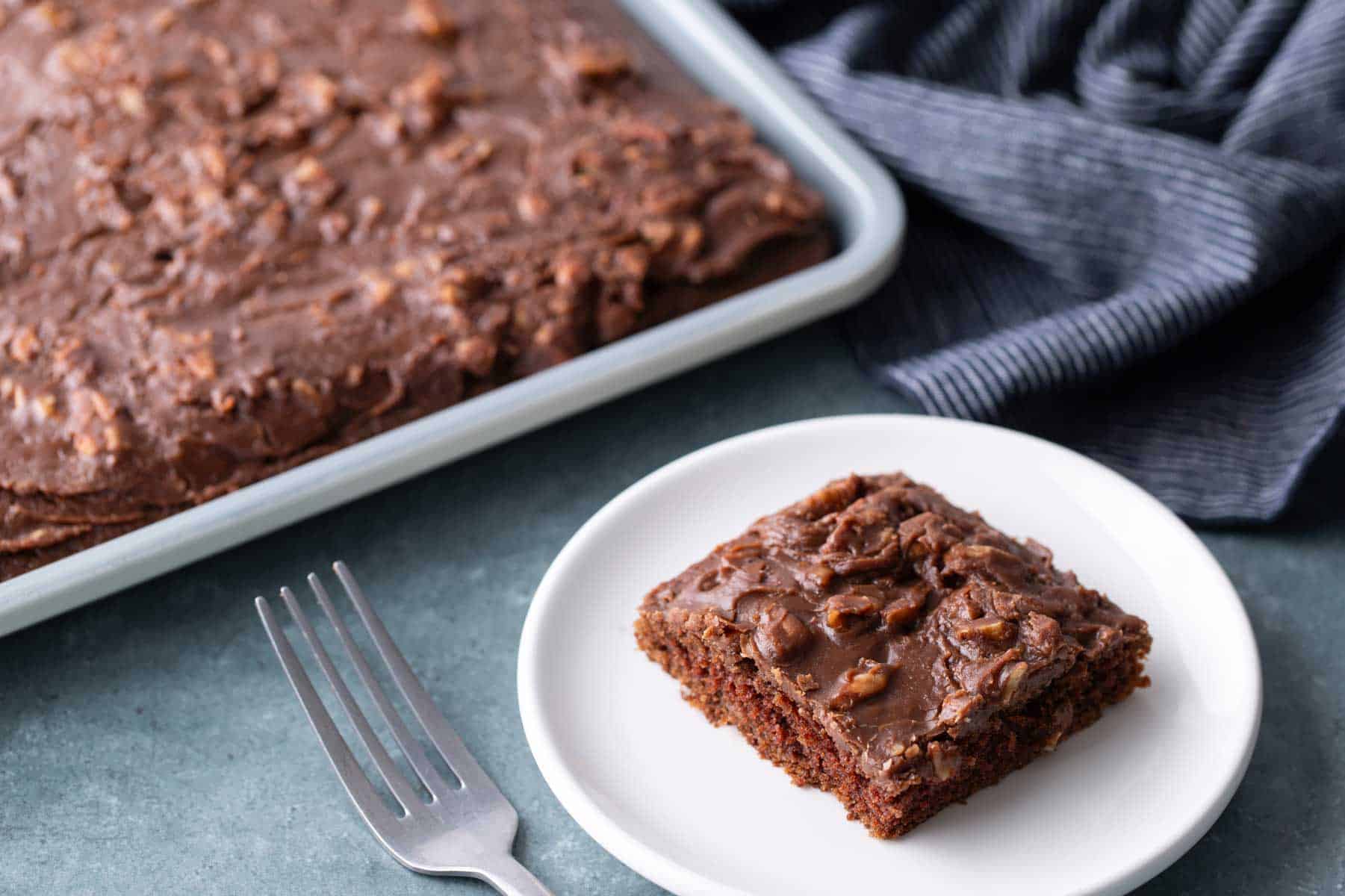 A chocolate sheet cake with icing on a baking tray, with a single square piece served on a white plate next to a fork.