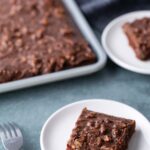A chocolate sheet cake with nuts on a baking tray, with two square pieces served on white plates and a fork nearby.