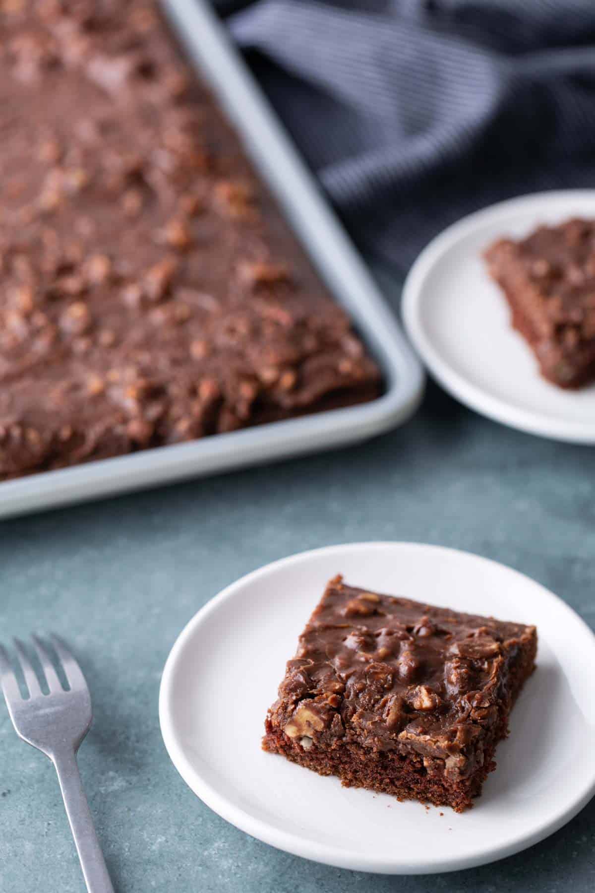 A chocolate sheet cake with nuts on a baking tray, with two square pieces served on white plates and a fork nearby.