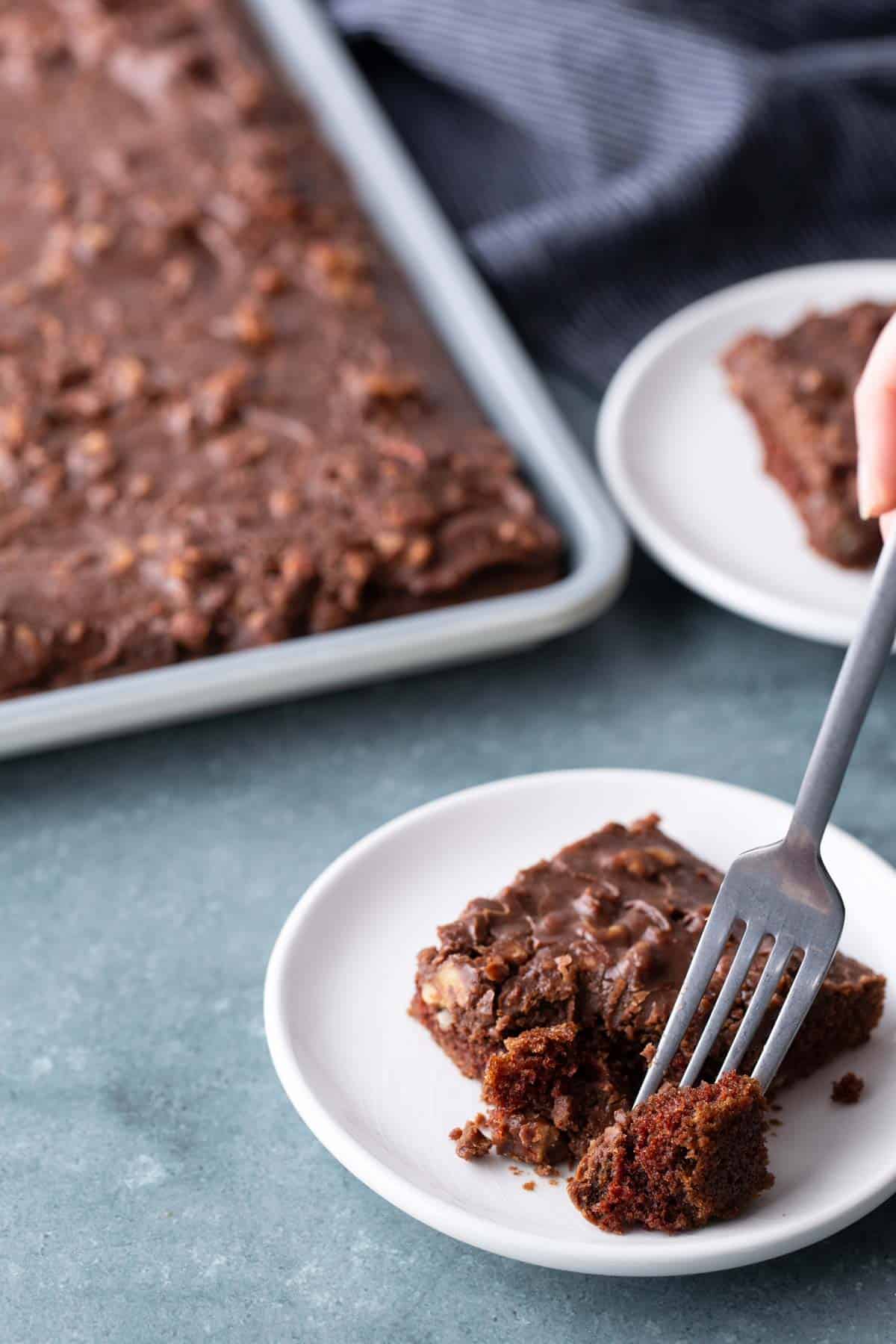A hand uses a fork to take a bite of chocolate sheet cake on a white plate, with a tray of cake and another plate in the background.