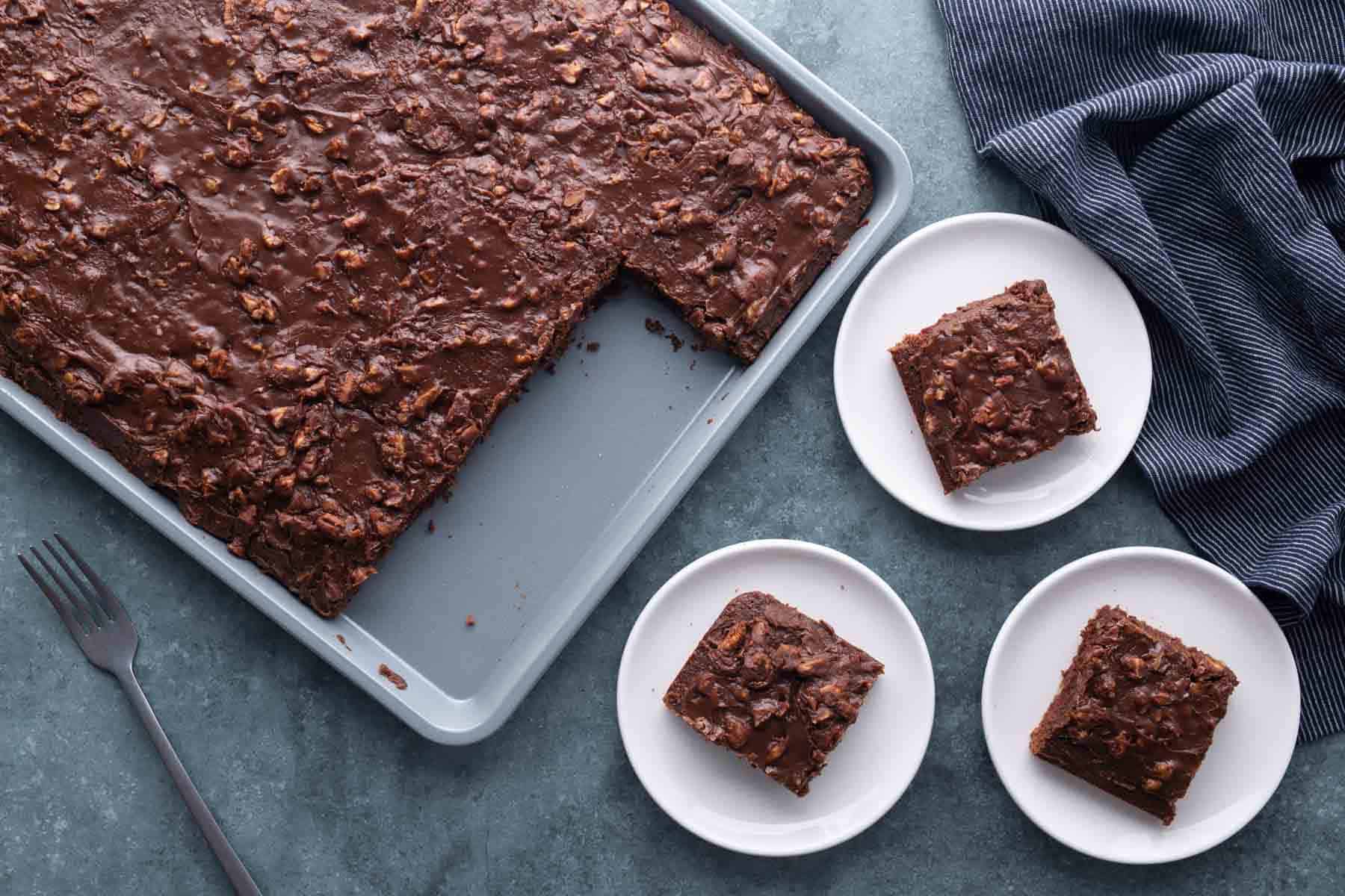 A tray of chocolate sheet cake with a piece missing is on a table, next to three plates each holding a square piece of the cake, with a fork and striped cloth nearby.