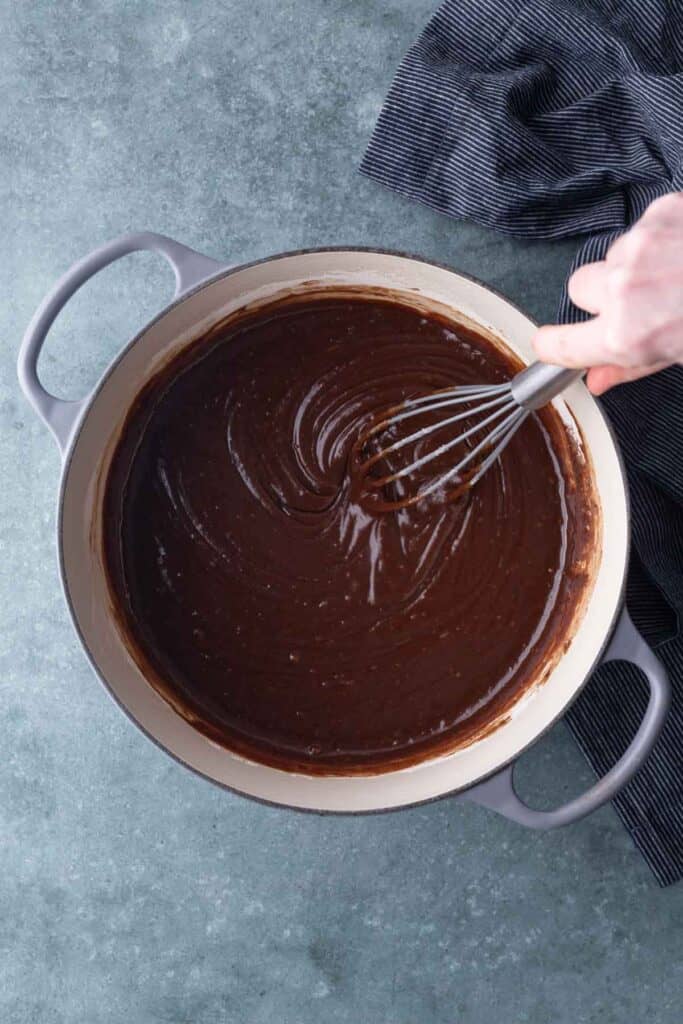 A hand uses a whisk to stir chocolate batter in a large gray pot on a gray countertop, with a striped cloth nearby.