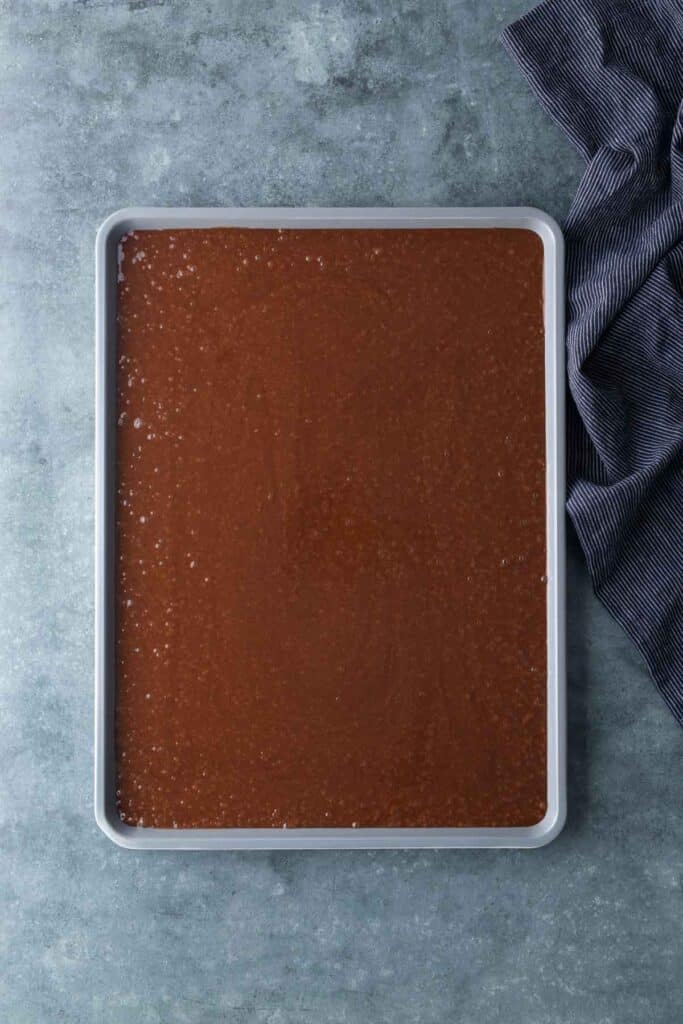 A rectangular baking pan filled with chocolate cake batter sits on a gray countertop next to a dark-striped cloth.