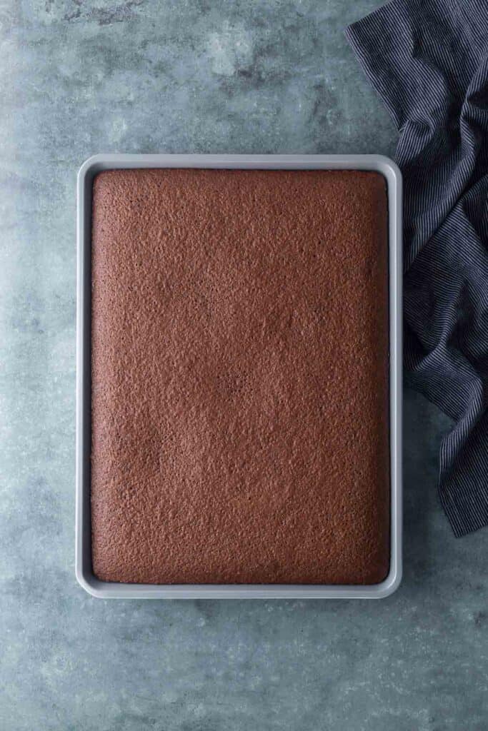 A rectangular chocolate sheet cake in a metal baking pan on a gray surface next to a dark striped cloth.
