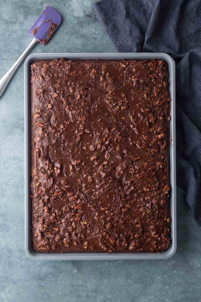 A rectangular baking pan filled with a chocolate dessert sits on a gray surface next to a blue spatula and a dark striped cloth.