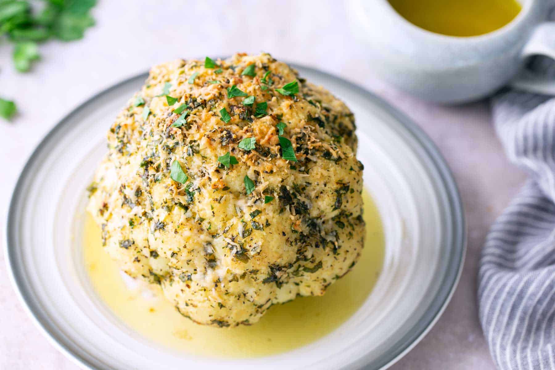 A whole roasted cauliflower topped with herbs and breadcrumbs sits on a plate with a pool of sauce, next to a cup of yellow liquid and a striped napkin.
