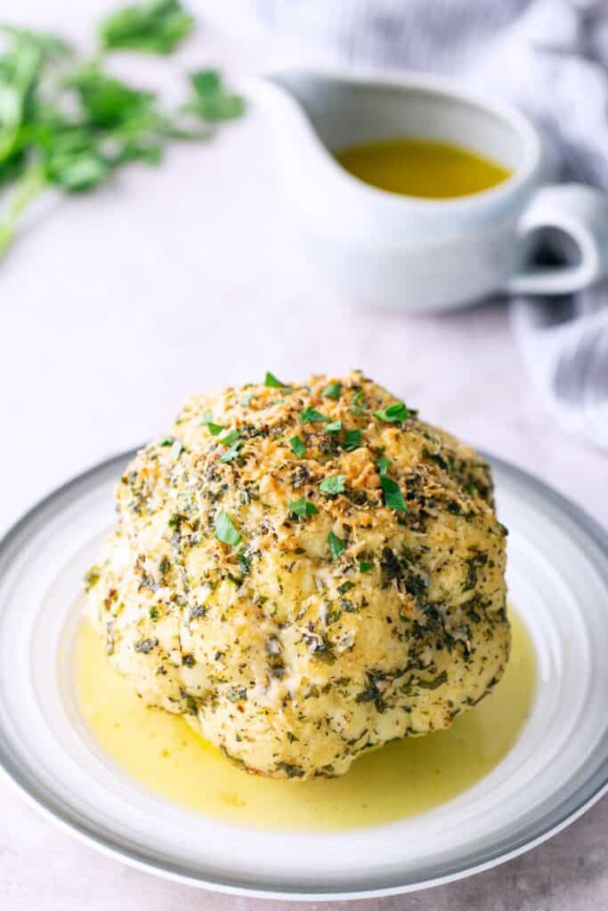 Whole Roasted Cauliflower Whole roasted cauliflower with herbs and seasoning, served on a plate with a pool of sauce; a jug of sauce and parsley in the background.