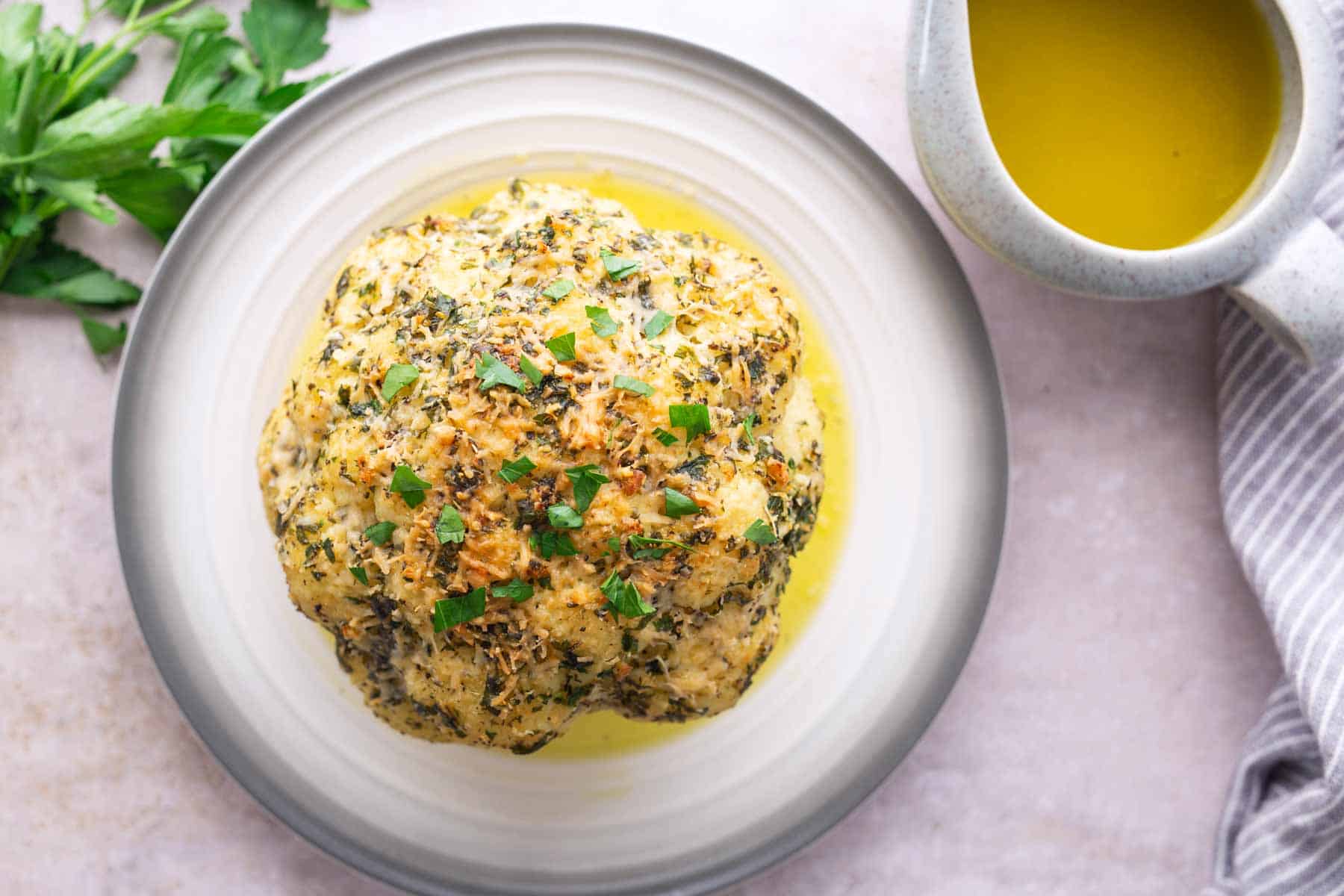 A whole roasted cauliflower topped with herbs and breadcrumbs on a plate, with a jug of sauce and fresh parsley on the side.