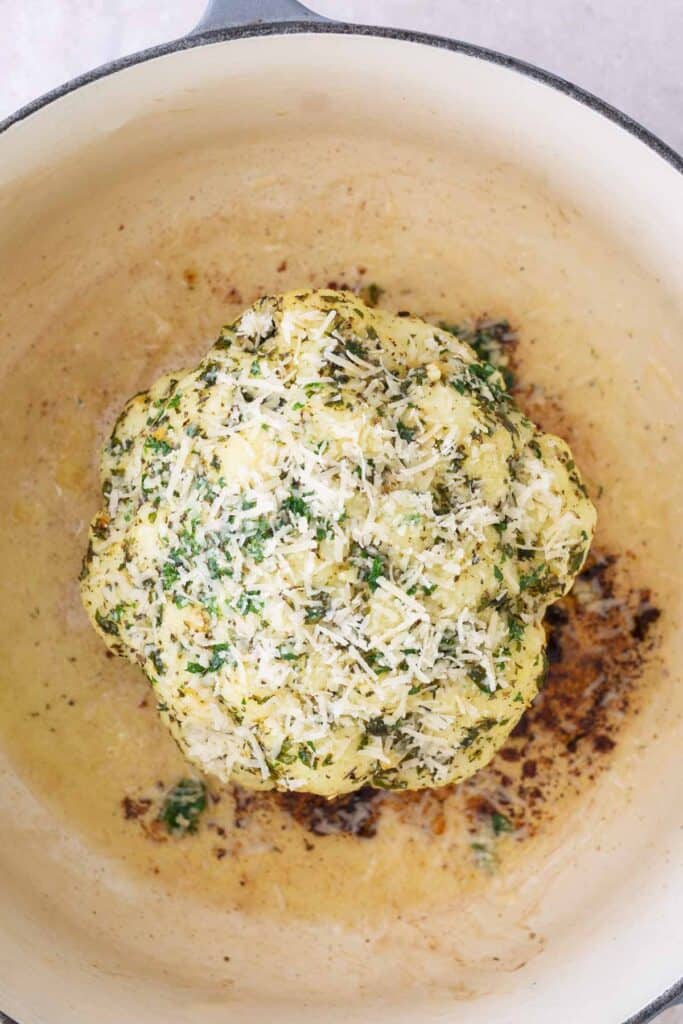 A whole roasted cauliflower topped with chopped herbs and grated cheese in a large, light-colored pot.