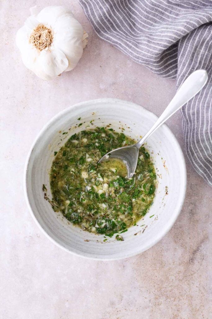 A white bowl with a spoon containing a herb and oil mixture sits on a light surface next to a bulb of garlic and a gray striped cloth.