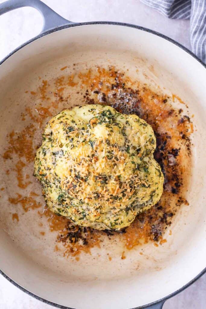 Whole roasted cauliflower coated with herbs and spices sits in a white Dutch oven, surrounded by browned bits and oil.