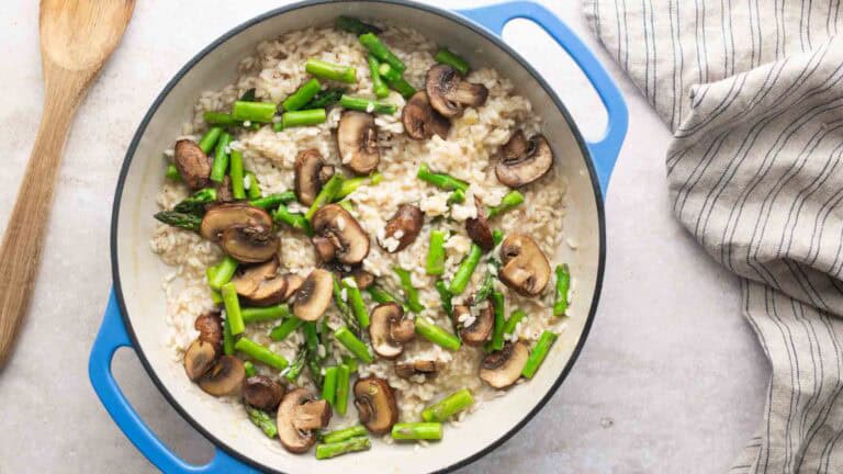 A blue pot filled with risotto, topped with sautéed mushrooms and asparagus, sits on a countertop next to a wooden spoon and a striped cloth.