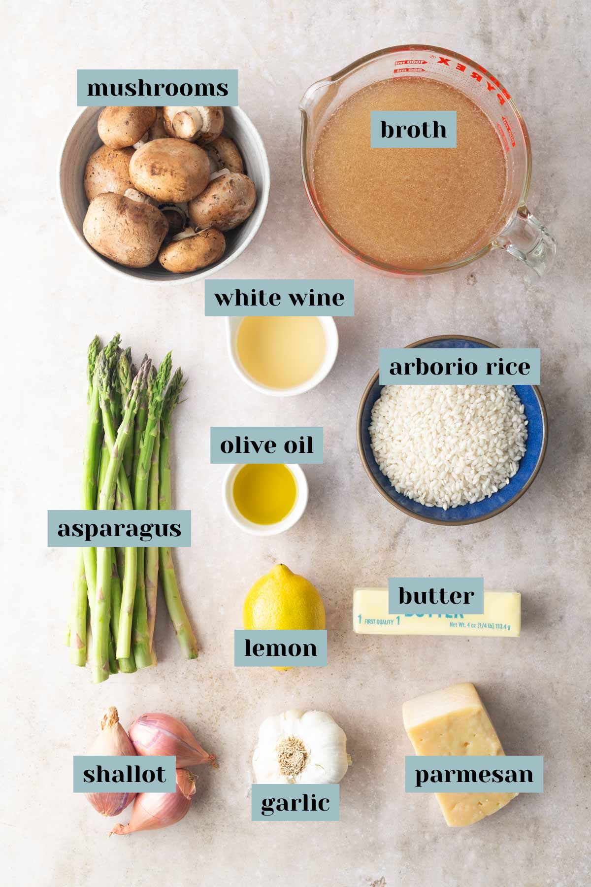 Ingredients for risotto including mushrooms, broth, white wine, asparagus, olive oil, arborio rice, lemon, butter, shallot, garlic, and parmesan, arranged on a light surface.