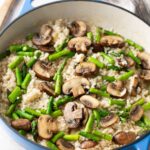 A blue pot filled with risotto, topped with sliced mushrooms and pieces of asparagus, sits on a light surface next to a wooden spoon and a striped cloth.
