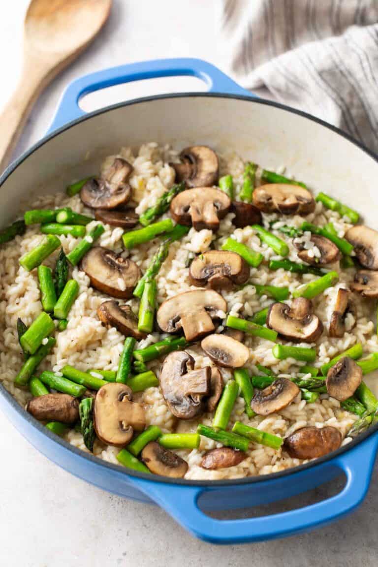 A blue pot filled with risotto, topped with sliced mushrooms and pieces of asparagus, sits on a light surface next to a wooden spoon and a striped cloth.