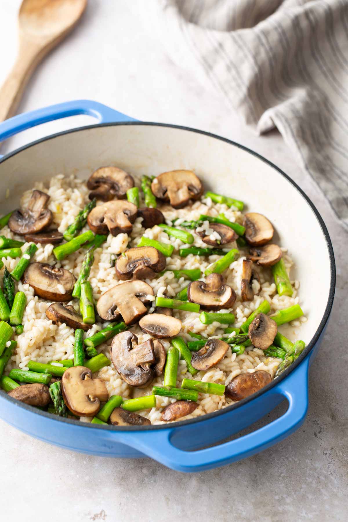 A blue casserole dish filled with cooked rice, sliced mushrooms, and chopped asparagus, with a wooden spoon and a striped cloth nearby.