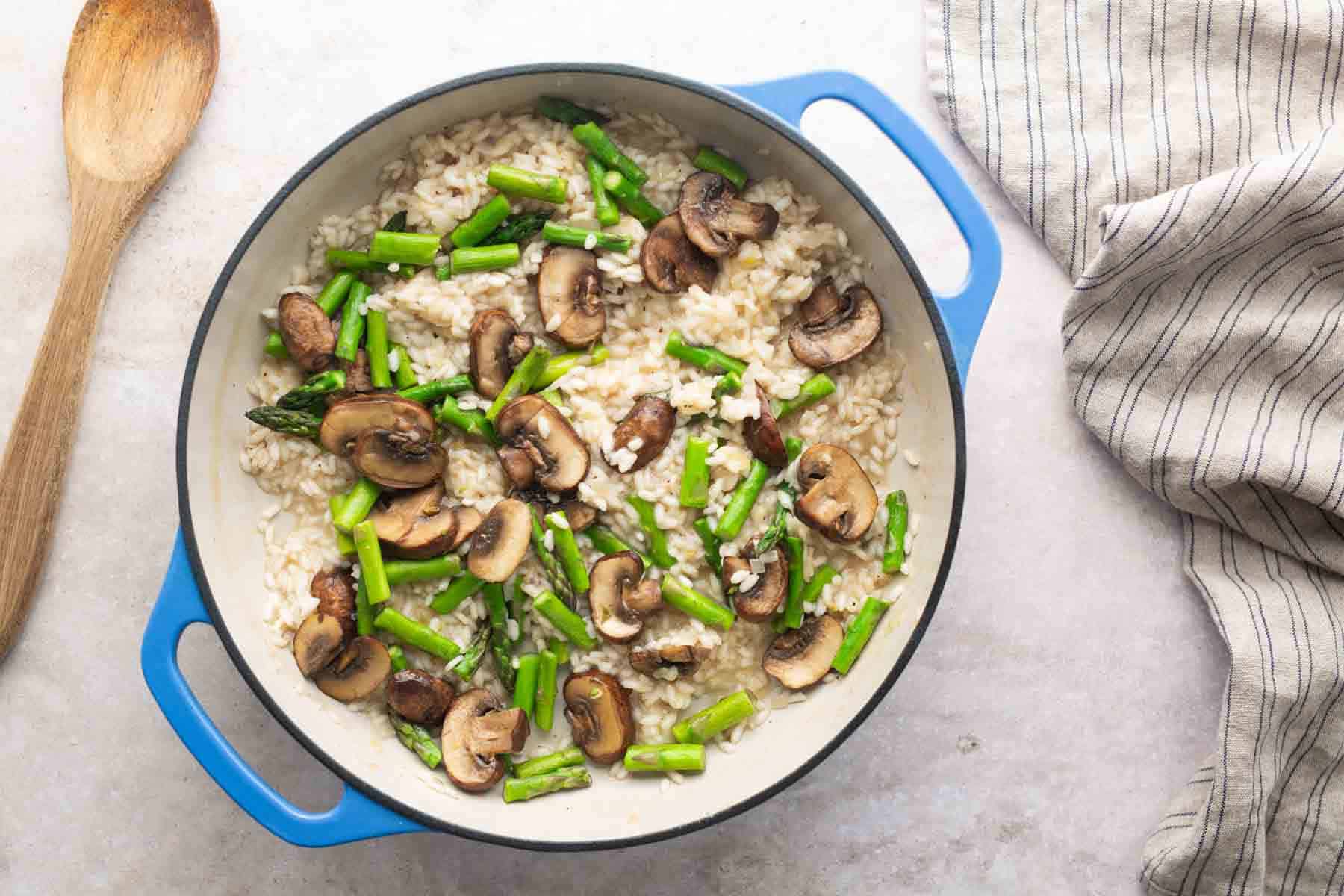 A blue pot filled with risotto topped with sliced mushrooms and asparagus, next to a wooden spoon and a striped cloth on a light surface.