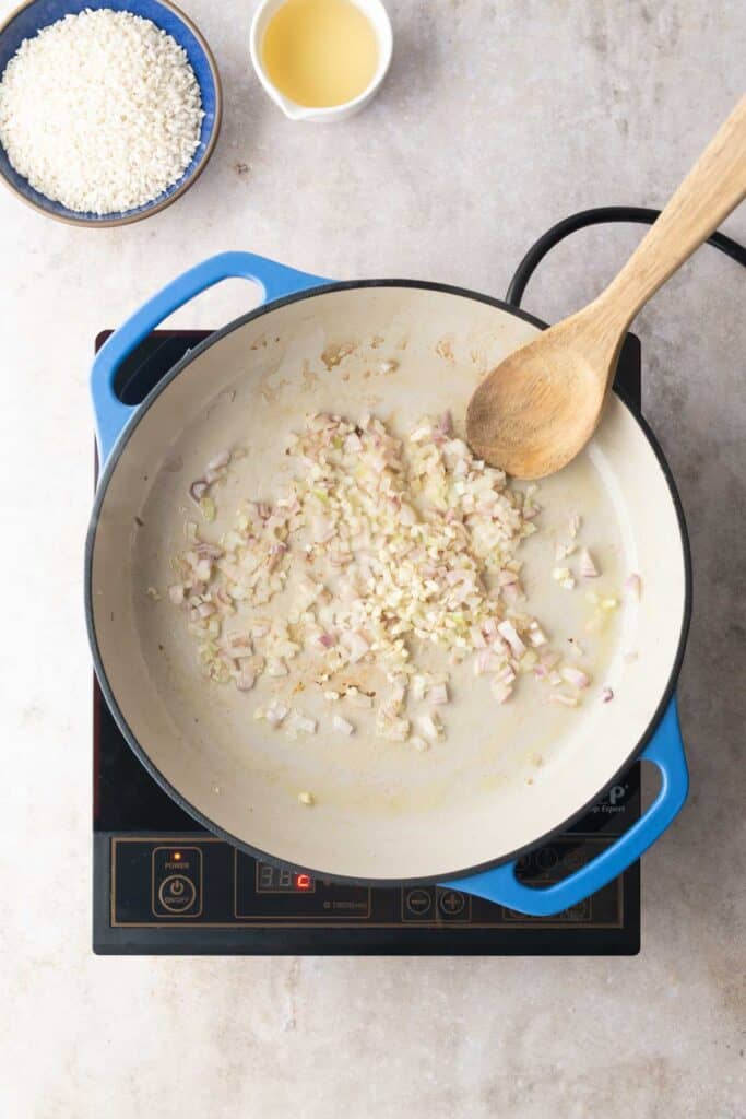 Overhead view of chopped onions and garlic sautéing in a large blue pot on an electric stovetop, with a wooden spoon, a bowl of rice, and a small cup of broth nearby.