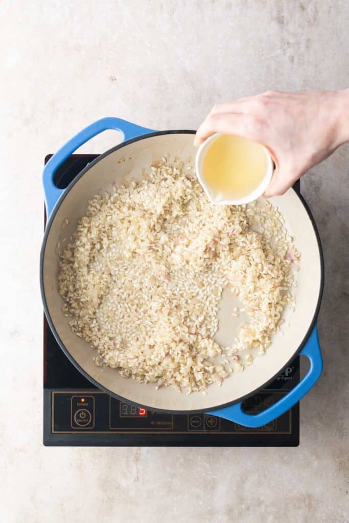 A hand pours liquid from a small cup into a pan of rice cooking on an electric stovetop.