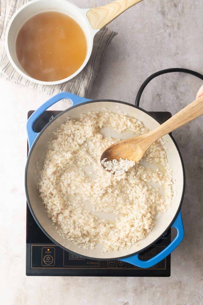 A wooden spoon stirs rice in a blue pot on an induction cooktop, with a saucepan of broth nearby on a striped cloth.