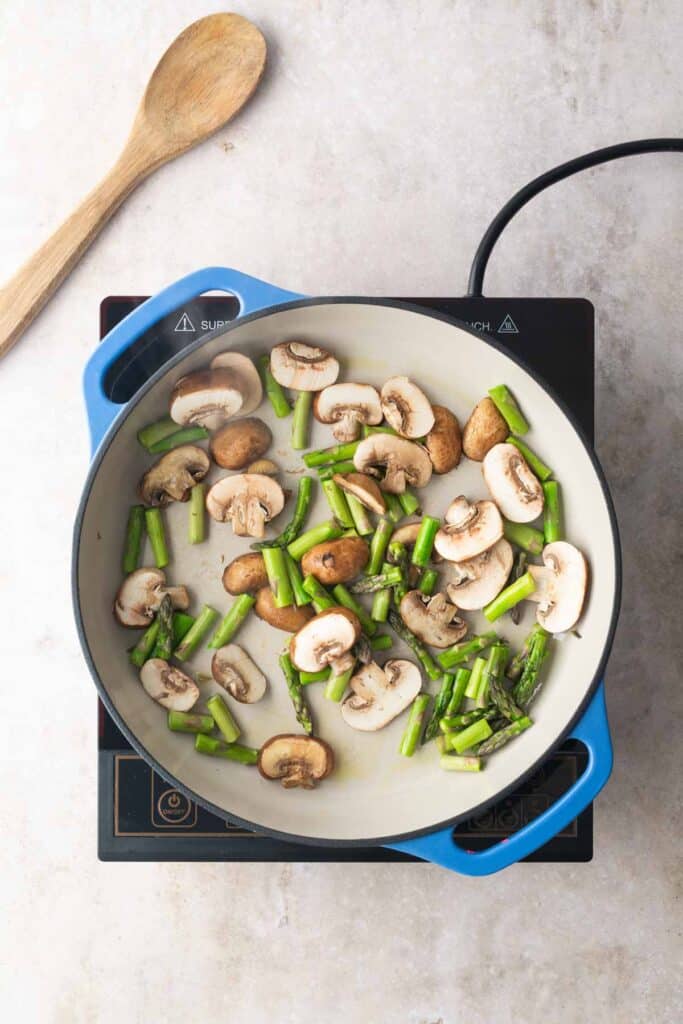 Sliced mushrooms and chopped asparagus cooking in a blue pan on an induction cooktop, with a wooden spoon nearby.