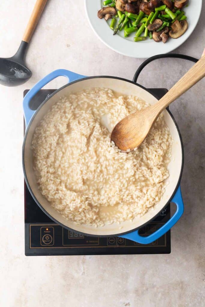 A pot of risotto cooking on an induction stovetop with a wooden spoon, next to a plate of sautéed mushrooms and asparagus.