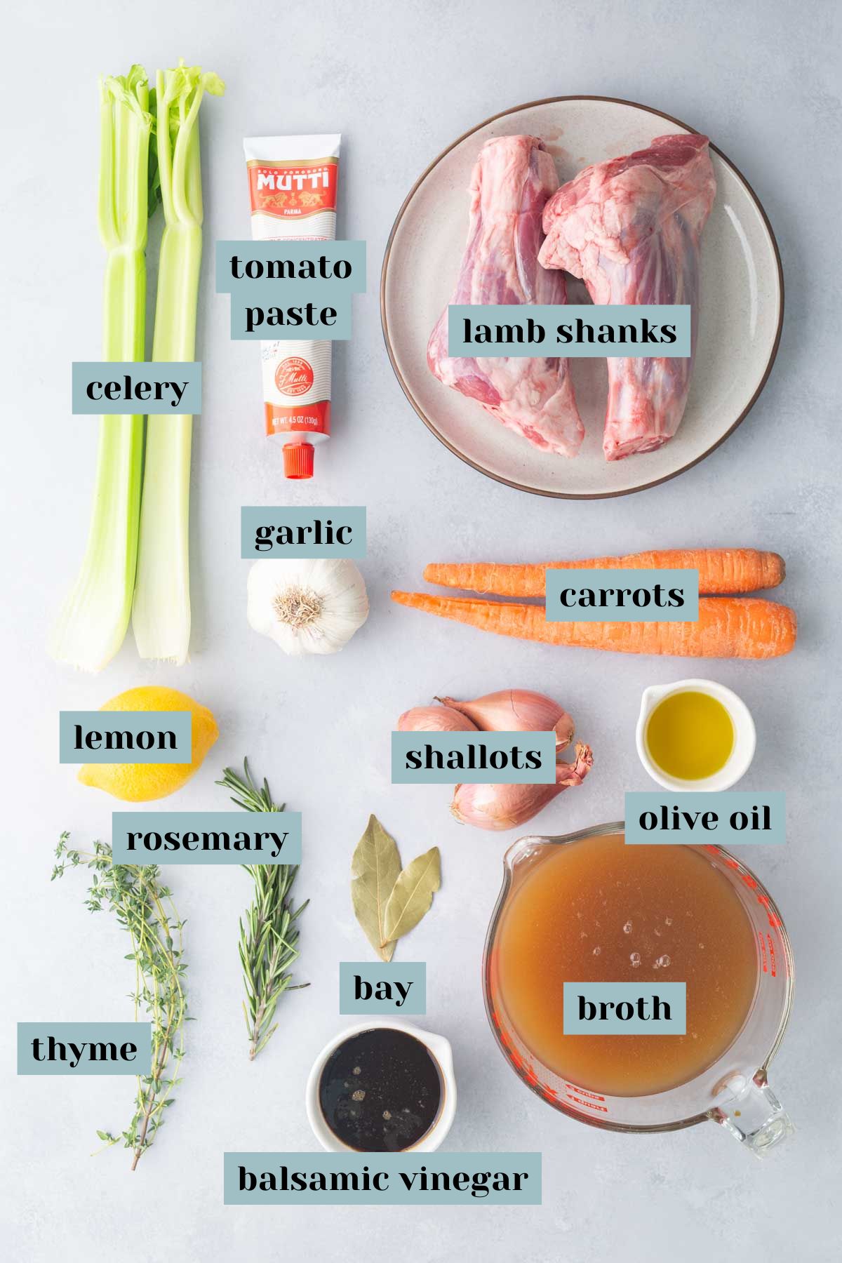 Labeled ingredients for lamb shanks laid out on a countertop, including lamb shanks, celery, tomato paste, garlic, carrots, lemon, olive oil, shallots, broth, herbs, bay leaves, and balsamic vinegar.