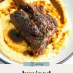 A braised lamb shank served on creamy mashed potatoes with gravy, garnished with herbs on a white plate.