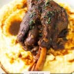 A braised lamb shank with sauce, garnished with herbs, served on a bed of creamy mashed potatoes on a white plate.