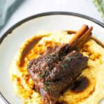 Braised lamb shank served on a bed of creamy mashed potatoes, garnished with fresh herbs and sauce on a white plate.