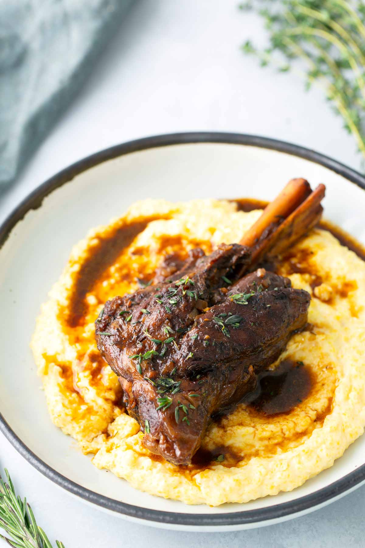 Braised lamb shank served on a bed of creamy mashed potatoes, garnished with fresh herbs and sauce on a white plate.