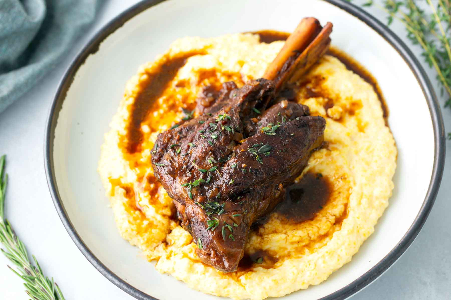 A cooked lamb shank with sauce sits on creamy polenta, garnished with fresh herbs, on a white plate.