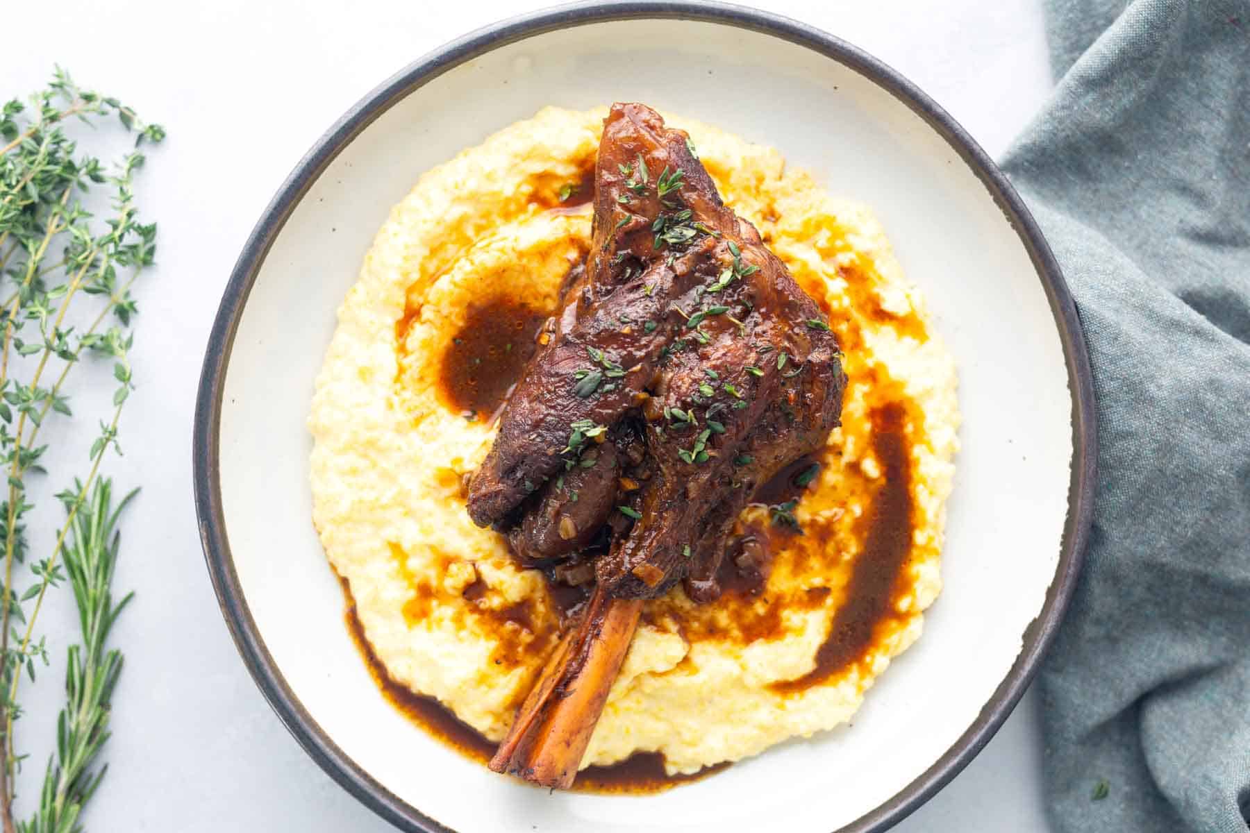 A braised lamb shank served on a bed of creamy polenta, garnished with fresh herbs and surrounded by sauce.