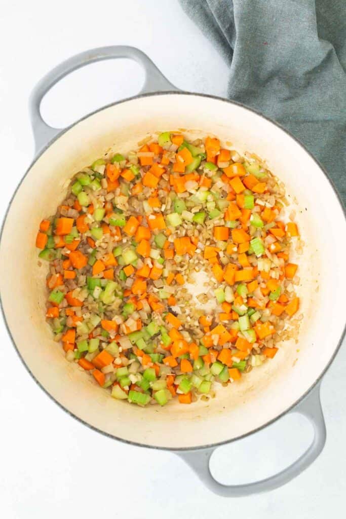 Diced carrots, celery, and onions sautéing in a large white pot, with a gray cloth in the background.