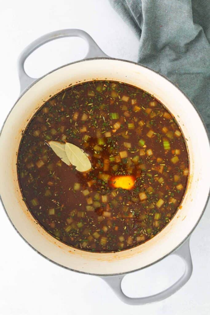 A large pot filled with dark broth containing diced vegetables, herbs, two bay leaves, and a pat of butter, with a cloth visible on the side.