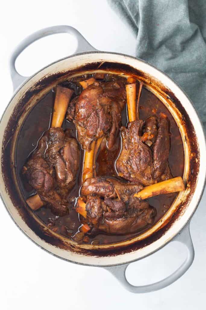 Four braised lamb shanks in a Dutch oven with a rich, dark sauce, viewed from above. A gray cloth is partially visible beside the pot.
