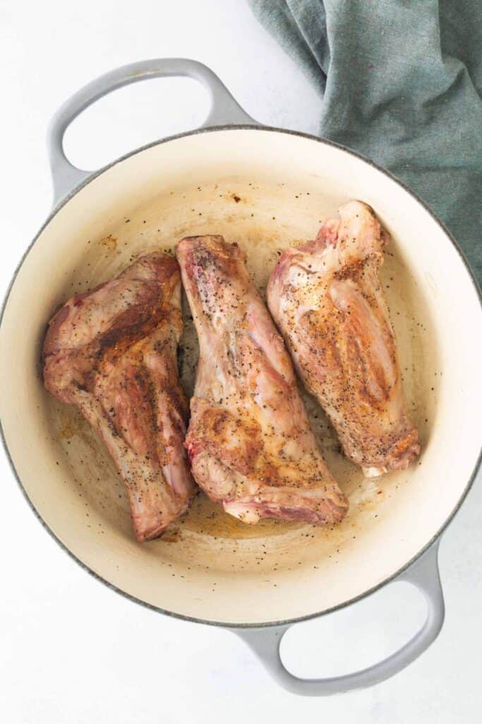 Three seasoned lamb shanks are browning in a white Dutch oven, with a green cloth partially visible in the upper right corner.