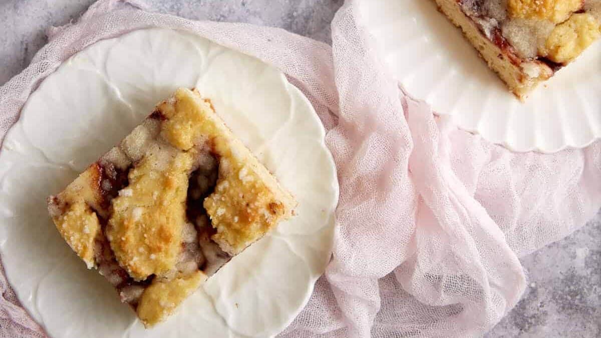 A piece of crumb-topped coffee cake sits on a white plate next to another partial piece, both on a light pink cloth over a gray surface.