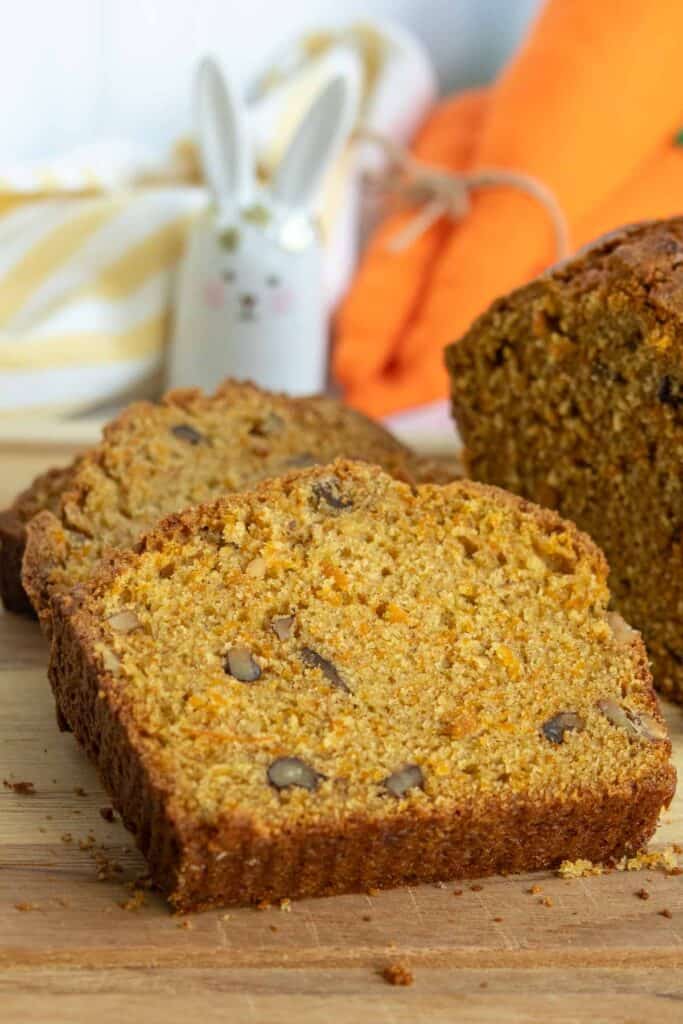 A close-up of sliced pumpkin bread with visible nuts on a wooden cutting board; a ceramic bunny and an orange cloth are in the background.