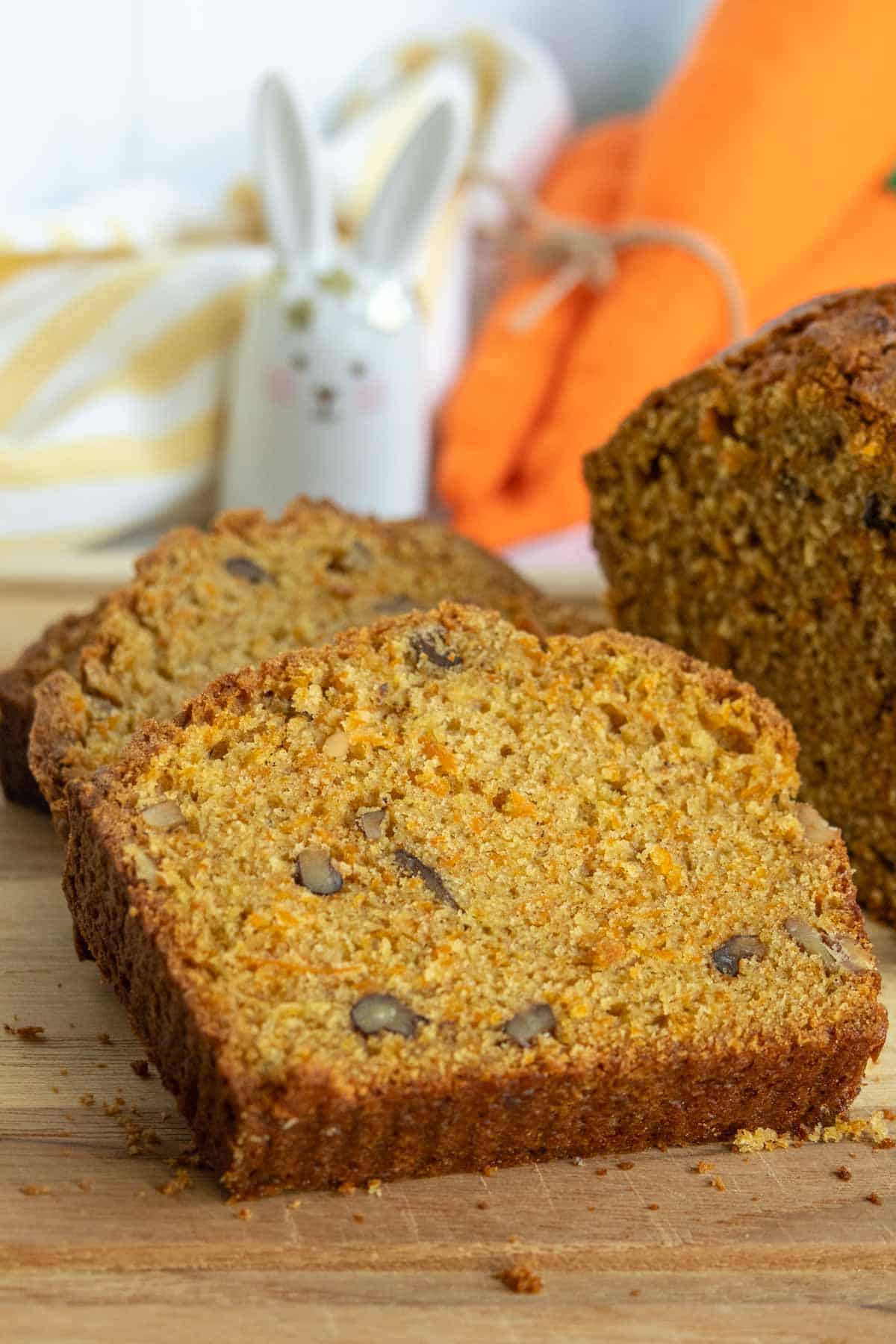 A close-up of sliced pumpkin bread with visible nuts on a wooden cutting board; a ceramic bunny and an orange cloth are in the background.