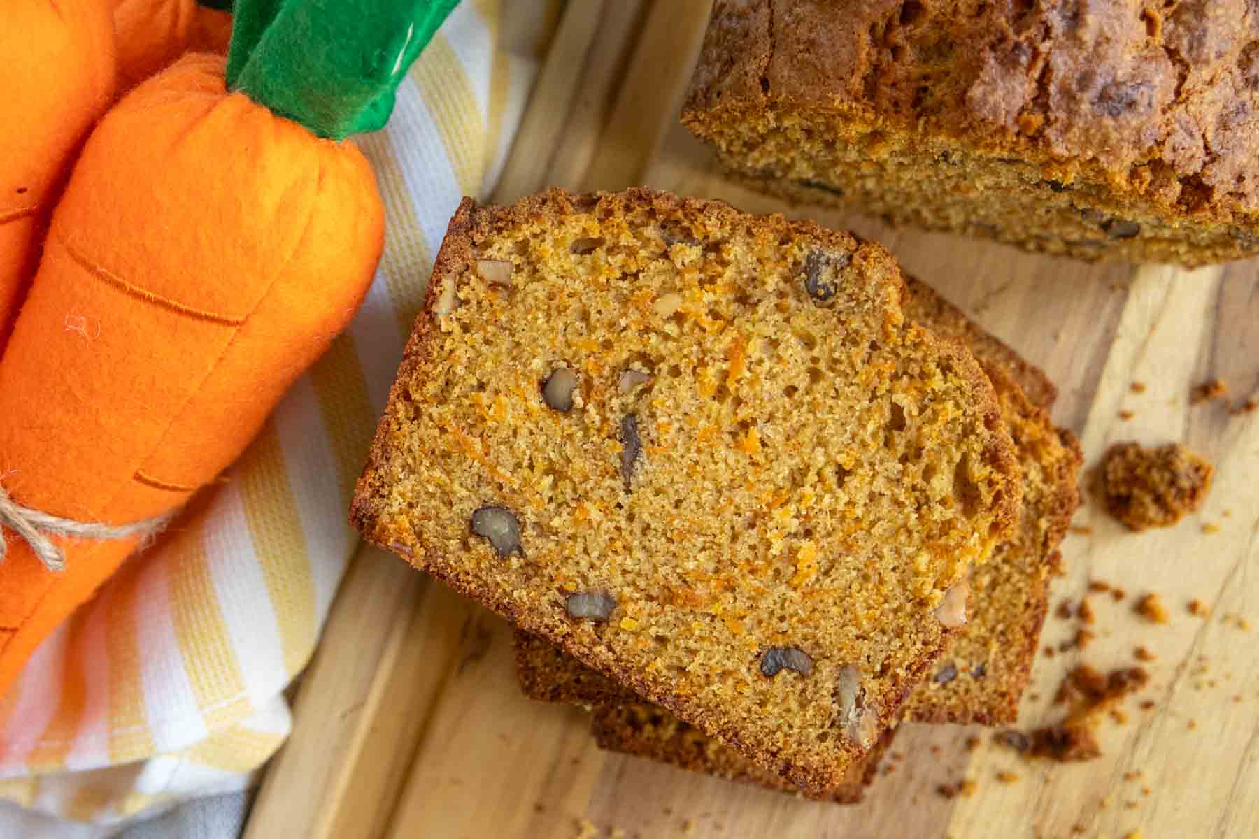 Two slices of carrot bread with nuts on a wooden board, next to a fabric carrot decoration and a striped cloth.