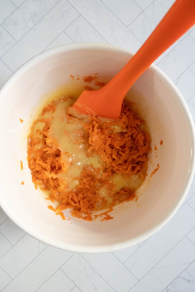 A white bowl contains grated carrots and liquid mixture being stirred with an orange spatula on a tiled surface.