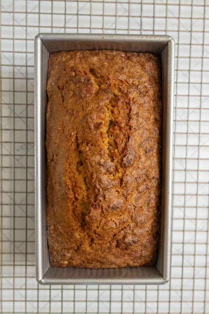 A loaf of banana bread in a metal baking pan sits on a wire cooling rack, viewed from above.
