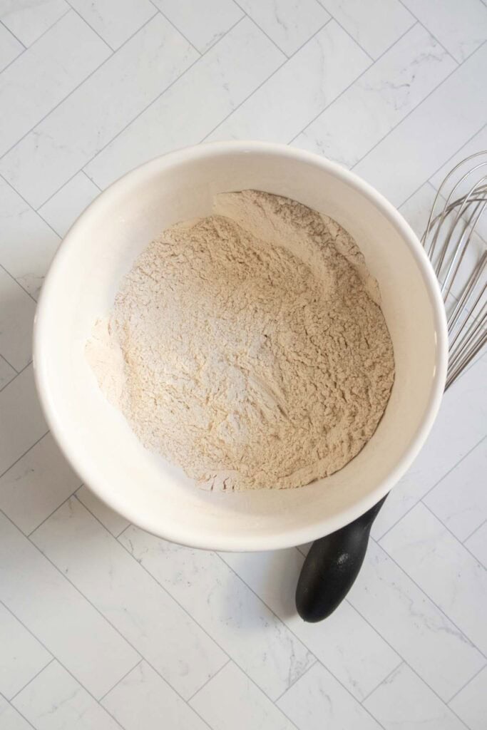 A large white mixing bowl filled with dry flour mixture sits on a light-colored tiled surface next to a metal whisk.