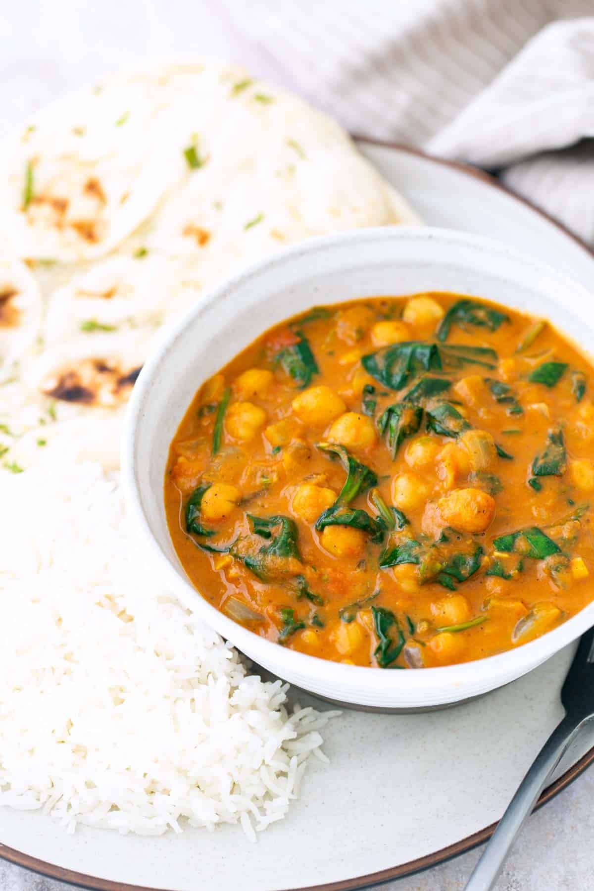 A bowl of chickpea and spinach curry with white rice and naan bread on a plate.