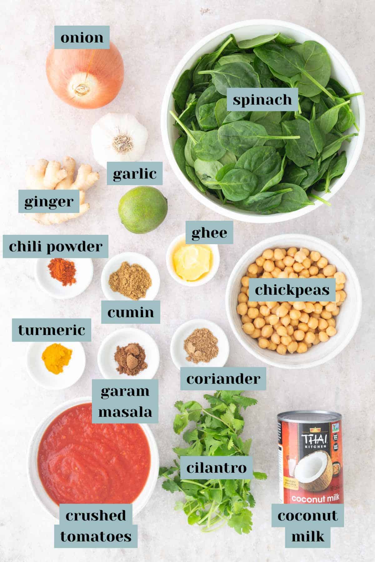 Labeled ingredients for a chickpea curry recipe, including onion, spinach, ginger, garlic, spices, ghee, chickpeas, cilantro, crushed tomatoes, and coconut milk on a countertop.