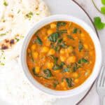 A bowl of chickpea and spinach curry served with white rice and garlic naan on a plate, with fresh cilantro leaves nearby.