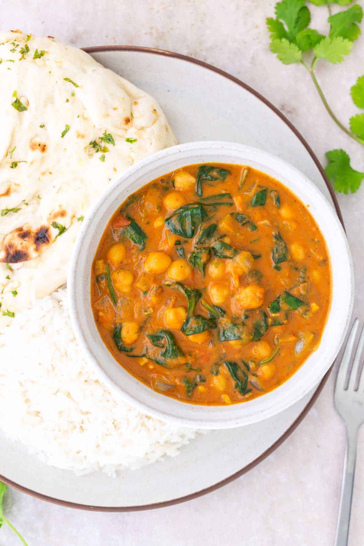 A bowl of chickpea and spinach curry served with white rice and garlic naan on a plate, with fresh cilantro leaves nearby.