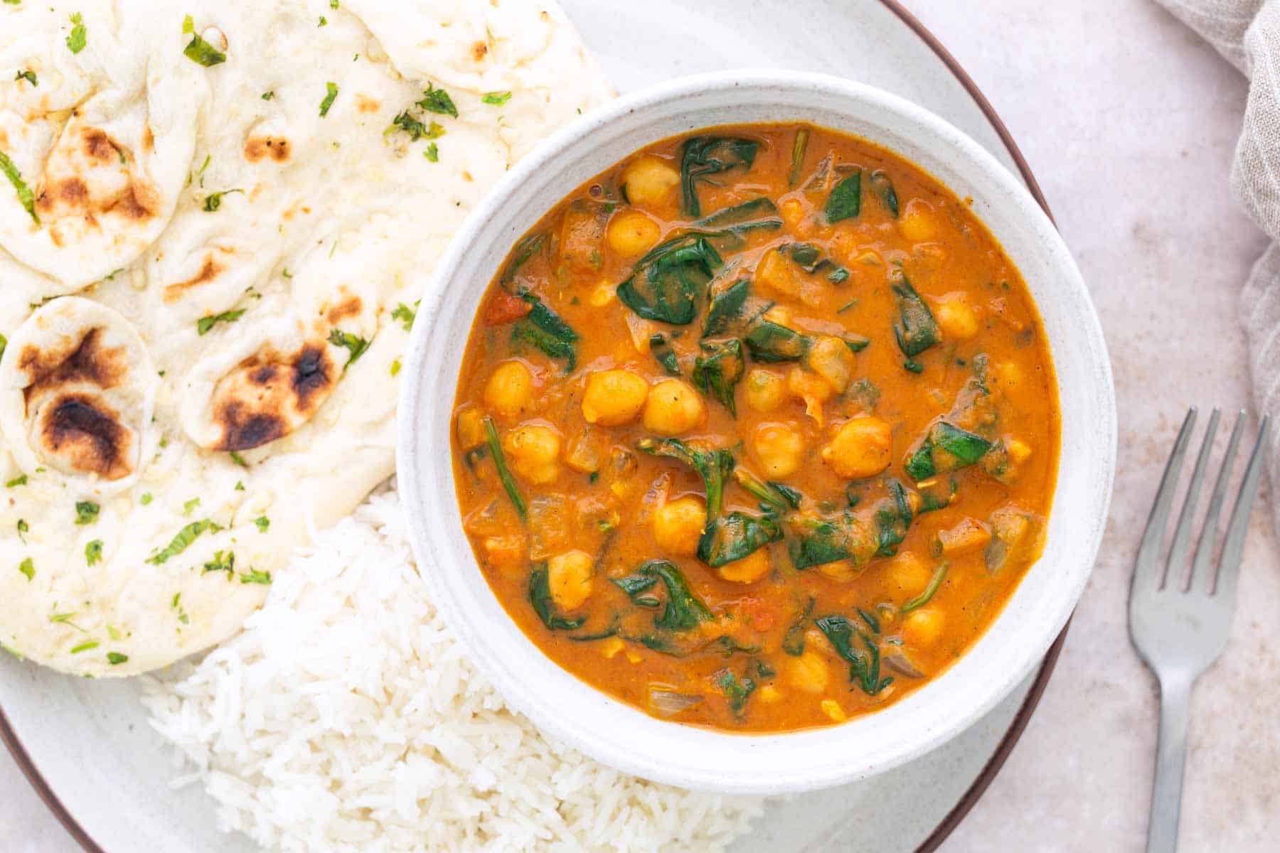 A bowl of chickpea and spinach curry with white rice and naan bread on a plate, next to a fork.
